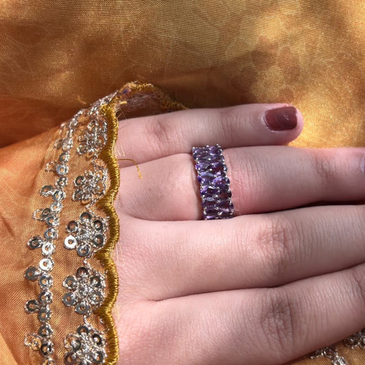 A close-up of a hand wearing a wide silver band ring set with purple stones, resting against a draped golden fabric background.