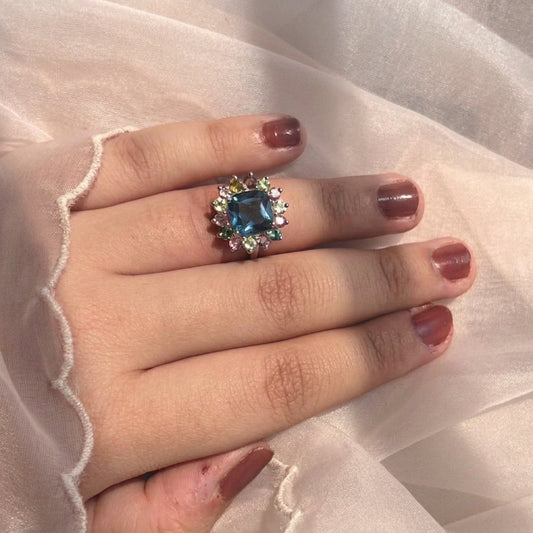 A close-up of a hand with mauve nail polish wearing a ring with a large blue gemstone surrounded by small clear stones, set against a background of sheer white fabric.