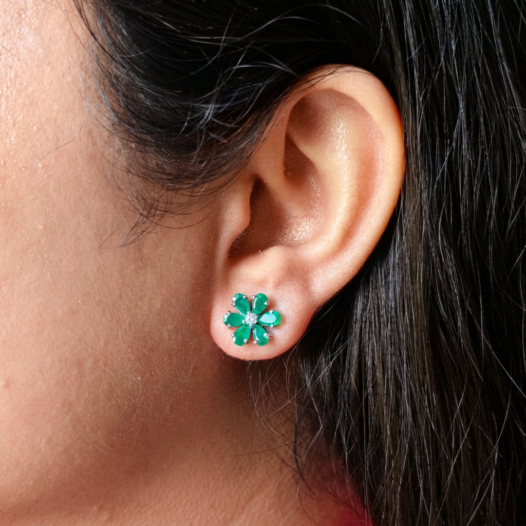 A close-up of a person's ear wearing a small flower-shaped stud earring with green stones and a clear center stone.