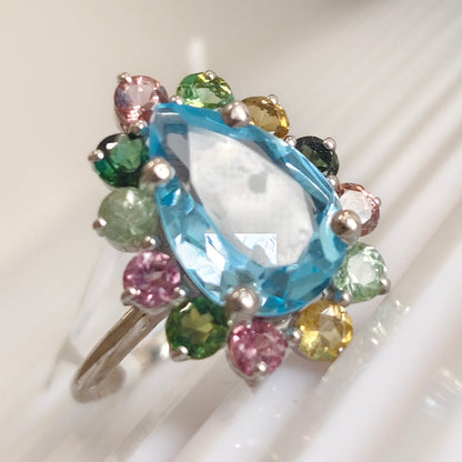 A multicolor gemstone ring featuring a large marquise-cut light blue central stone surrounded by a halo of small round-cut pink, green, yellow, and black stones in silver settings, displayed on a white ribbed surface.