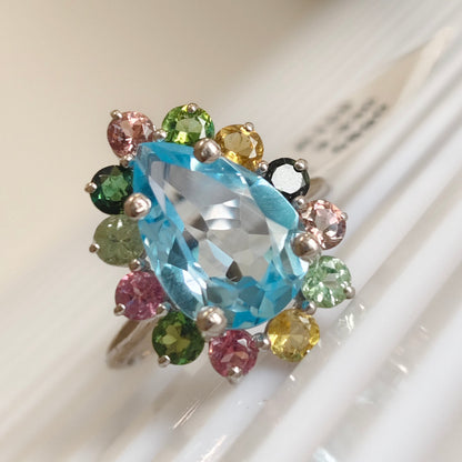 A silver ring featuring a large, faceted light blue marquise-cut center stone surrounded by a cluster of small, colorful round gemstones in pink, green, yellow, and black, resting on a white ribbed surface.