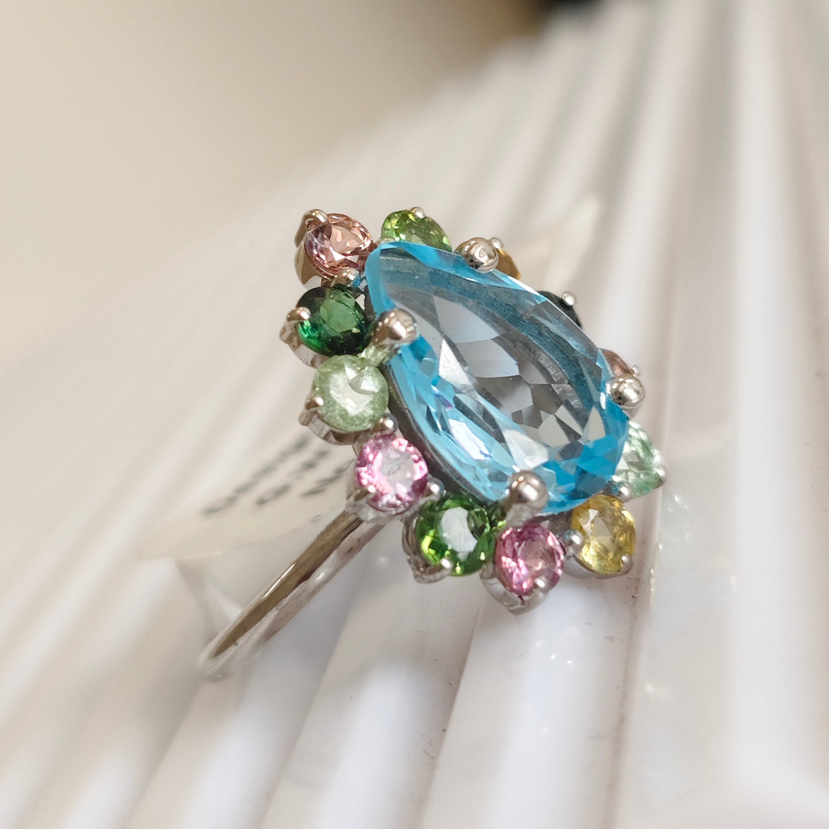 A silver ring featuring a large pear-cut light blue gemstone surrounded by a cluster of small multicolored round gemstones, displayed on a white textured surface.