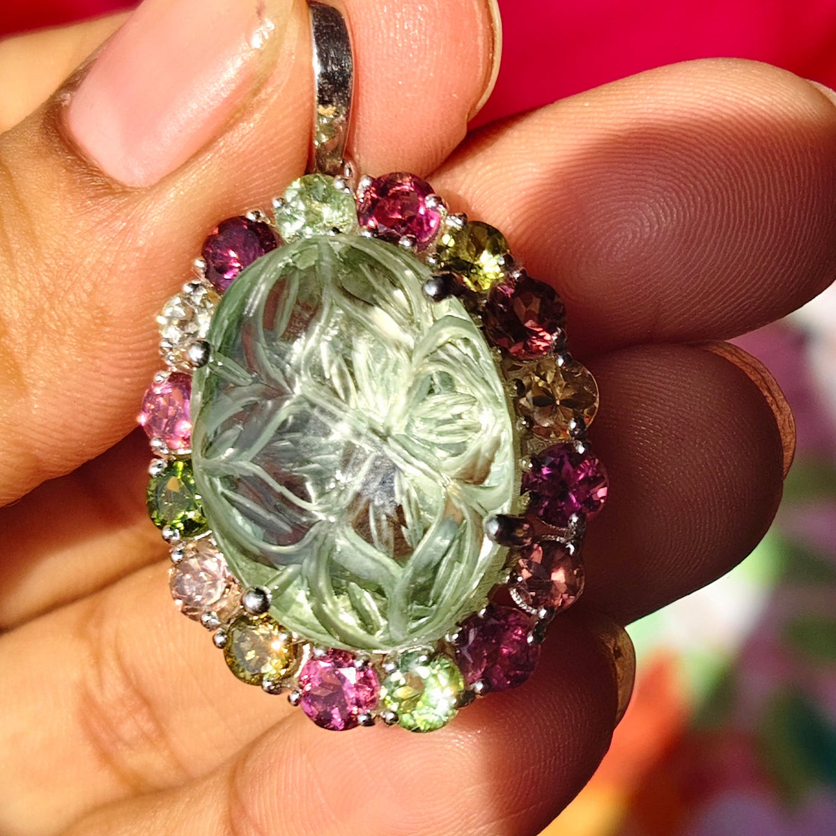 A hand holding a round pendant featuring a carved pale green stone surrounded by a border of small pink, green, and yellow rhinestones.