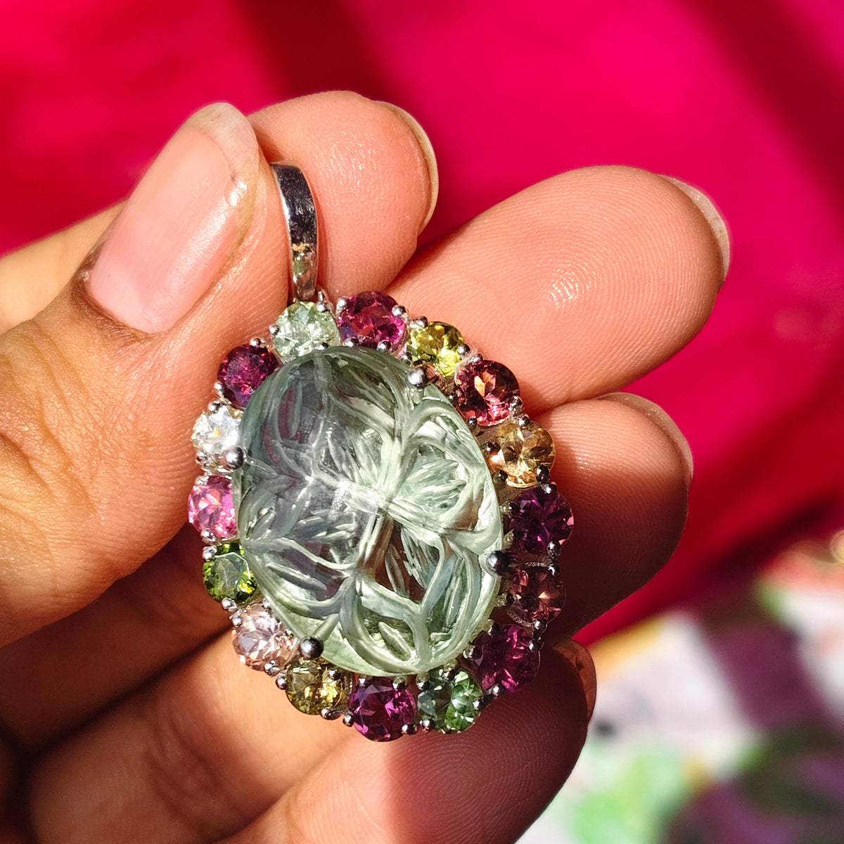 A hand holding a silver-toned oval pendant featuring a large clear faceted central stone surrounded by a halo of small multicolored gemstones, with a bright pink fabric background.