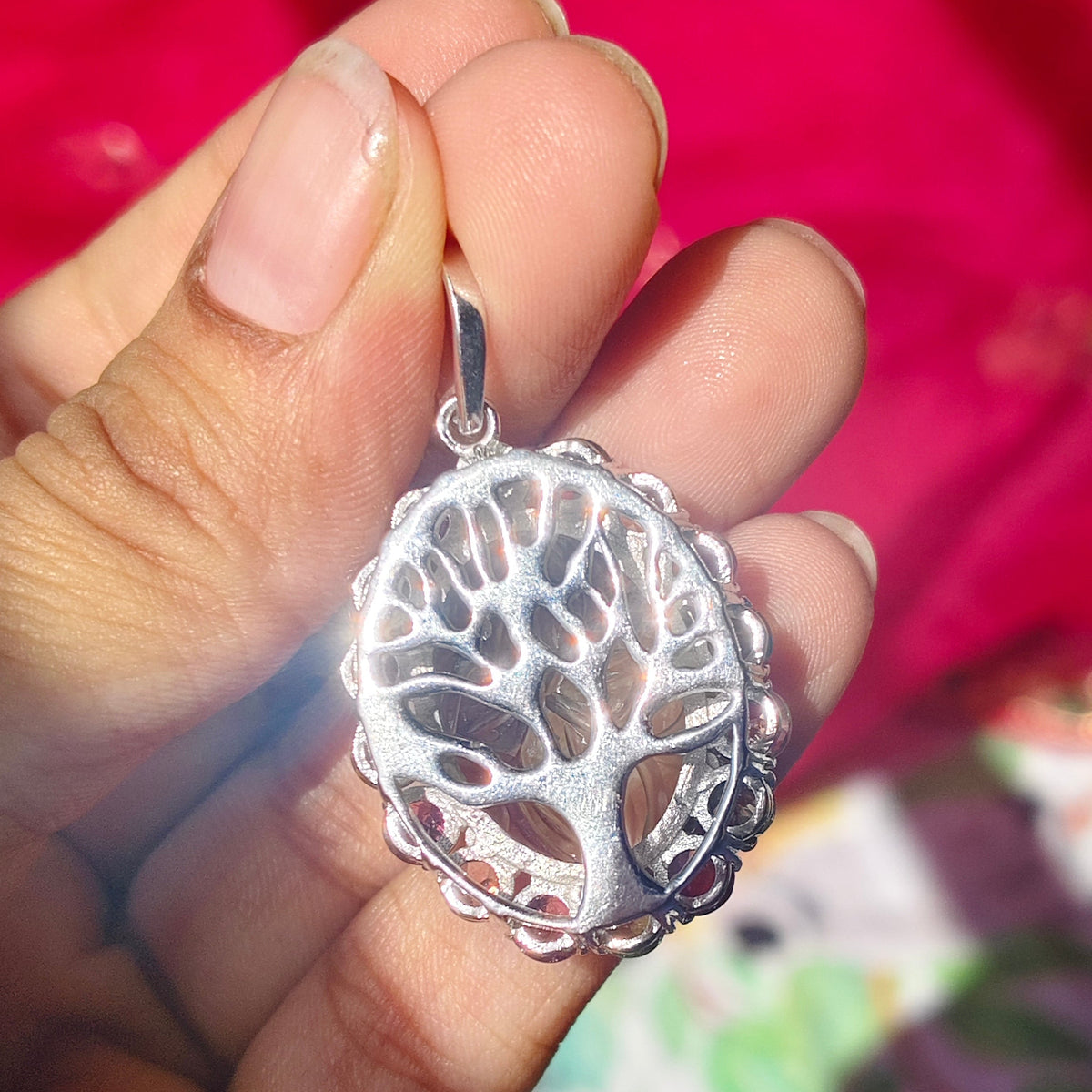 A hand holding a silver pendant with an intricate tree of life design, set against a vibrant pink and floral background.