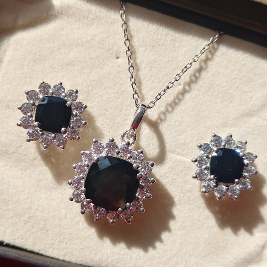 Silver-tone jewelry set with a pendant necklace and matching stud earrings, featuring dark oval center stones surrounded by clear rhinestone-like accents, displayed on a cream jewelry box.