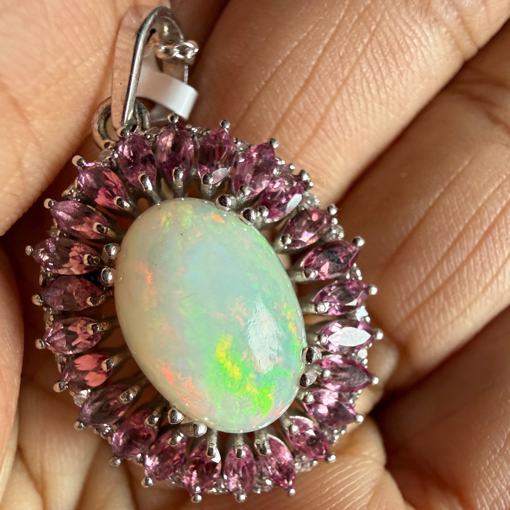 A close-up of a hand holding a silver halo ring featuring a central white opal with iridescent green and pink flashes, surrounded by a border of marquise-cut pink gemstones.