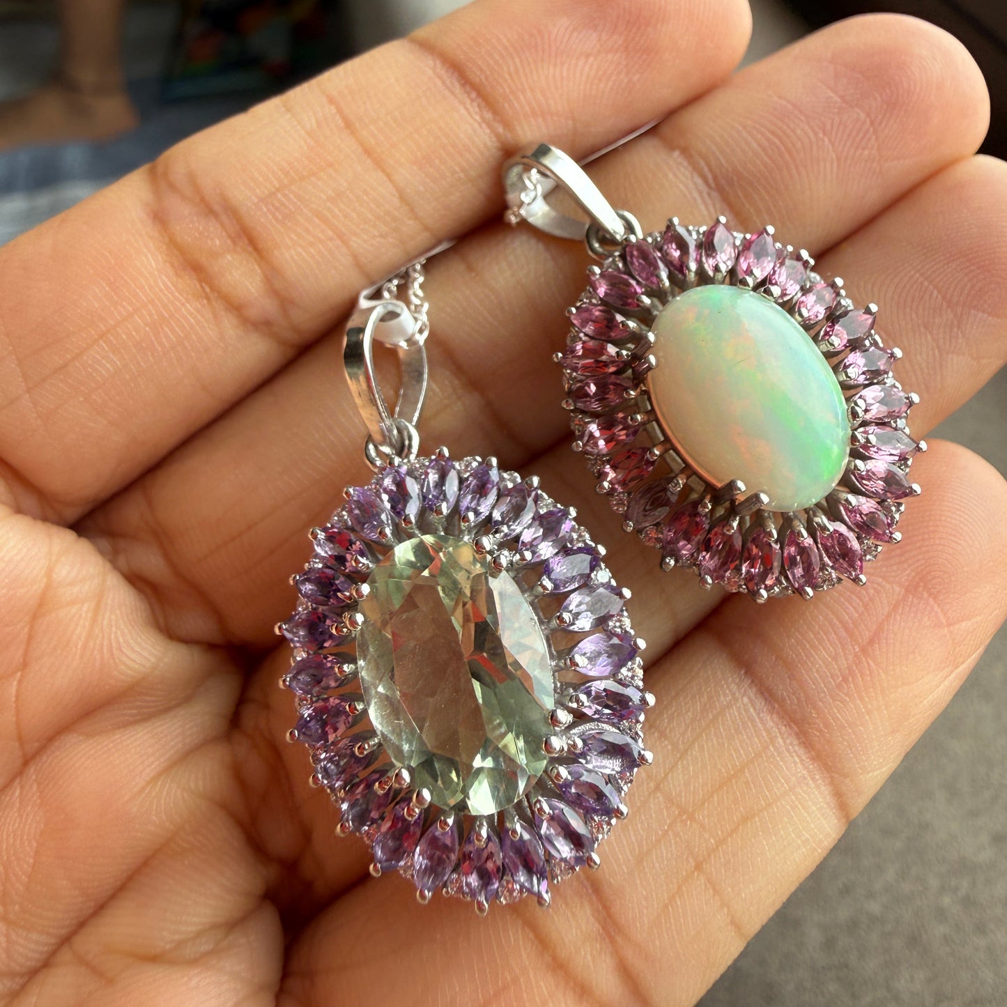 A pair of silver-toned dangle earrings with oval-cut iridescent opal centers surrounded by halo clusters of pink stones, held in a person's hand.