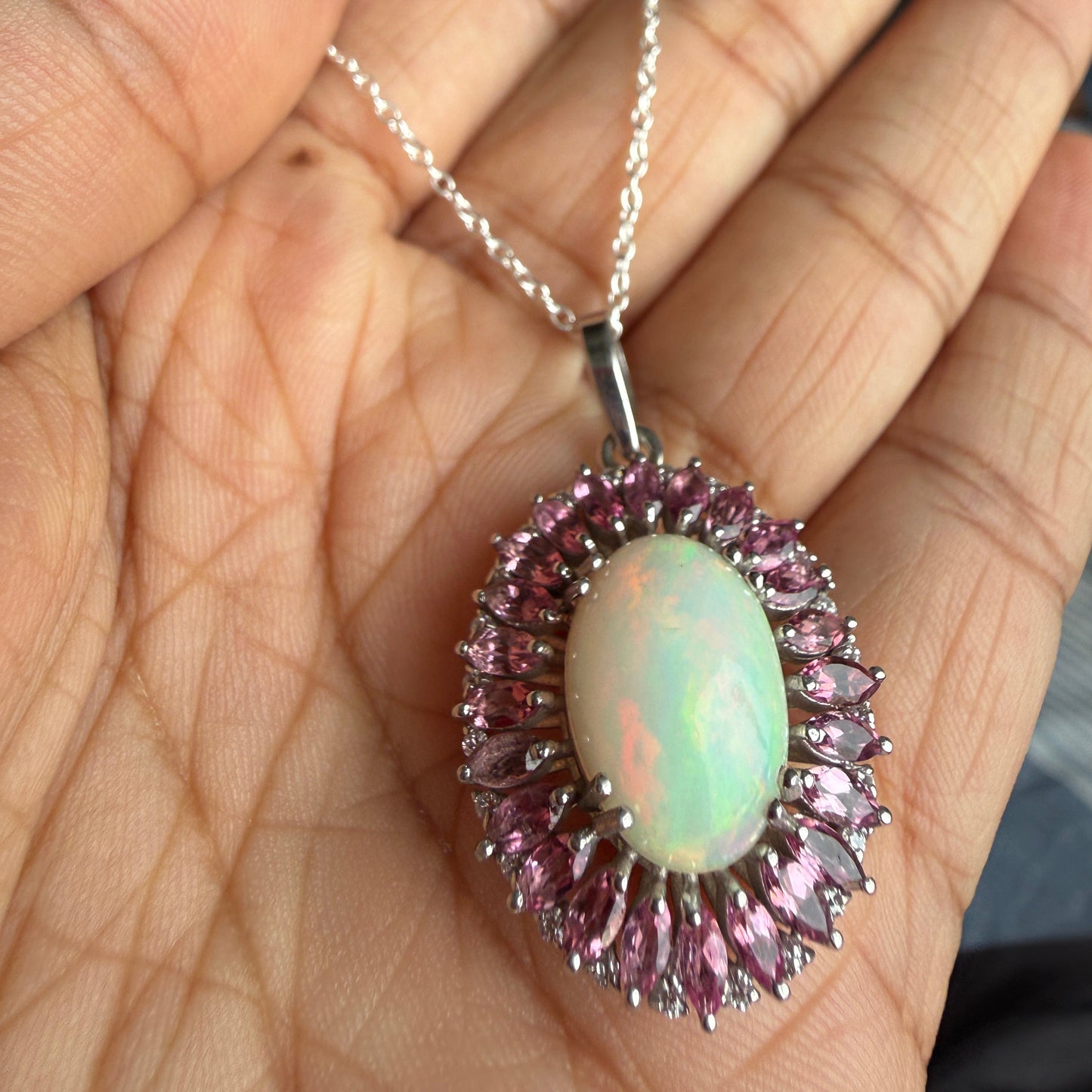 A silver pendant necklace with an oval iridescent opal center and surrounding pink gemstones, held in a person's hand.