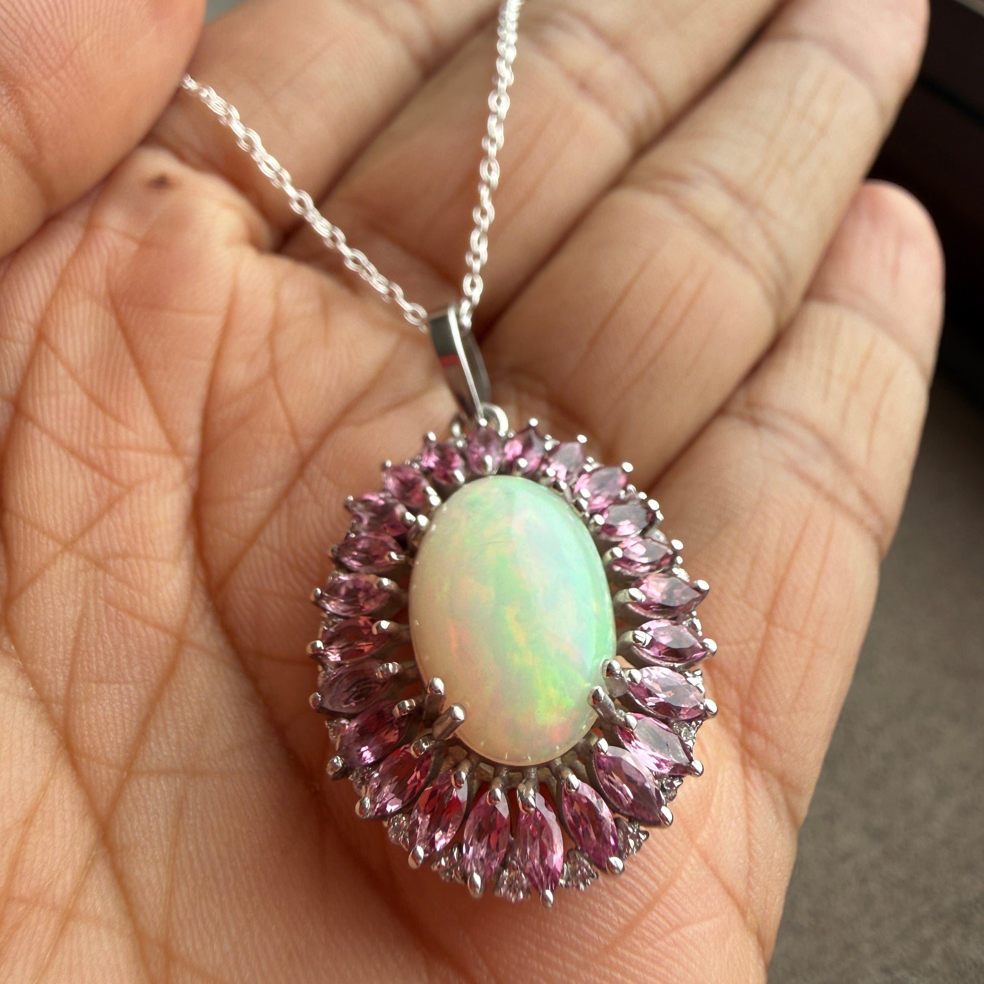 A silver pendant necklace with an oval iridescent white opal center and surrounding pink marquise-cut stones, held in a person's hand.
