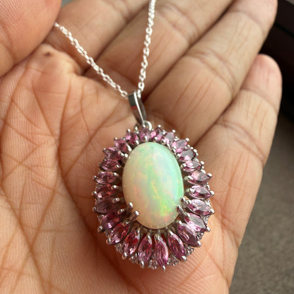A silver pendant necklace with an oval iridescent white opal center and surrounding pink marquise-cut stones, held in a person's hand.