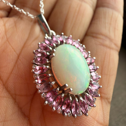 A close-up of a silver pendant necklace held in a hand. The pendant features a central oval cabochon opal with a pale green iridescent sheen, surrounded by a halo of marquise-cut pink stones and a border of small round clear stones.