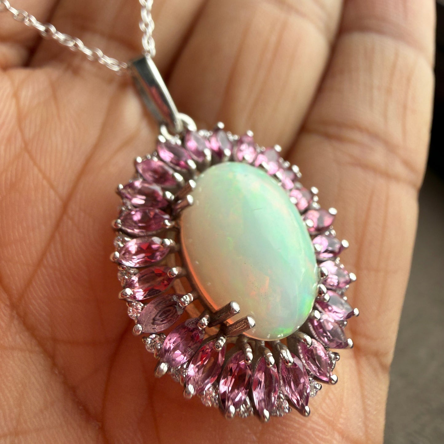 A close-up of a silver pendant necklace held in a hand. The pendant features a central oval cabochon opal with a pale green and white iridescent sheen, surrounded by a halo of marquise-cut pink stones in a silver setting, with a silver chain visible on the left.