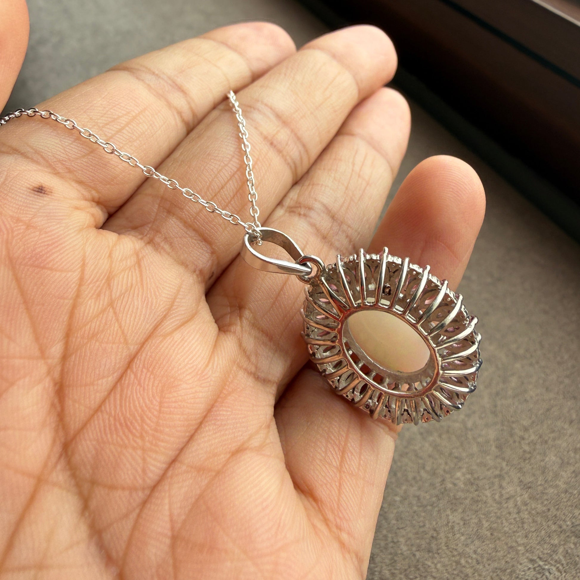 A silver-toned pendant necklace with a round beige stone and a decorative sunburst halo, held in a person's hand.