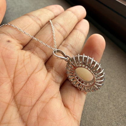 A silver-toned pendant necklace with a round beige stone and a decorative sunburst halo, held in a person's hand.