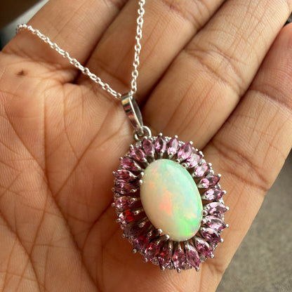 A close-up of a hand holding a silver pendant necklace with a central iridescent white opal surrounded by a halo of pink gemstones, with a silver chain draped across the fingers.