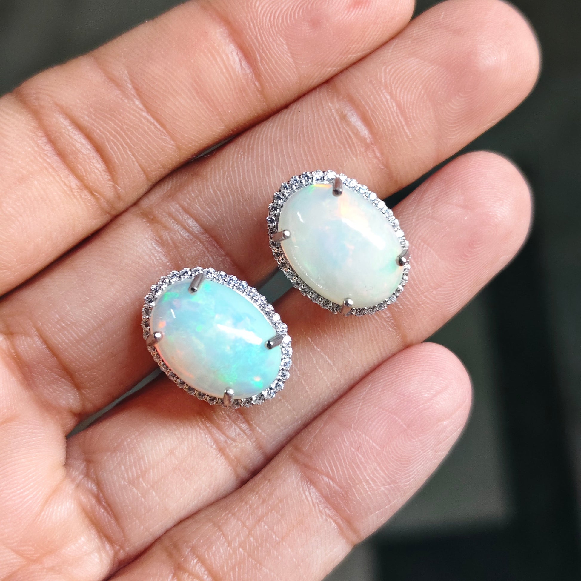 A pair of oval stud earrings with milky white iridescent opal-like centers and silver-toned halo settings, shown resting on a person's fingers against a dark blurred background.