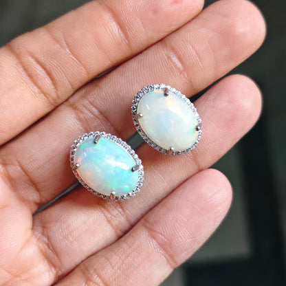 A pair of oval stud earrings with milky white iridescent opal-like centers and silver-toned halo settings, shown resting on a person's fingers against a dark blurred background.