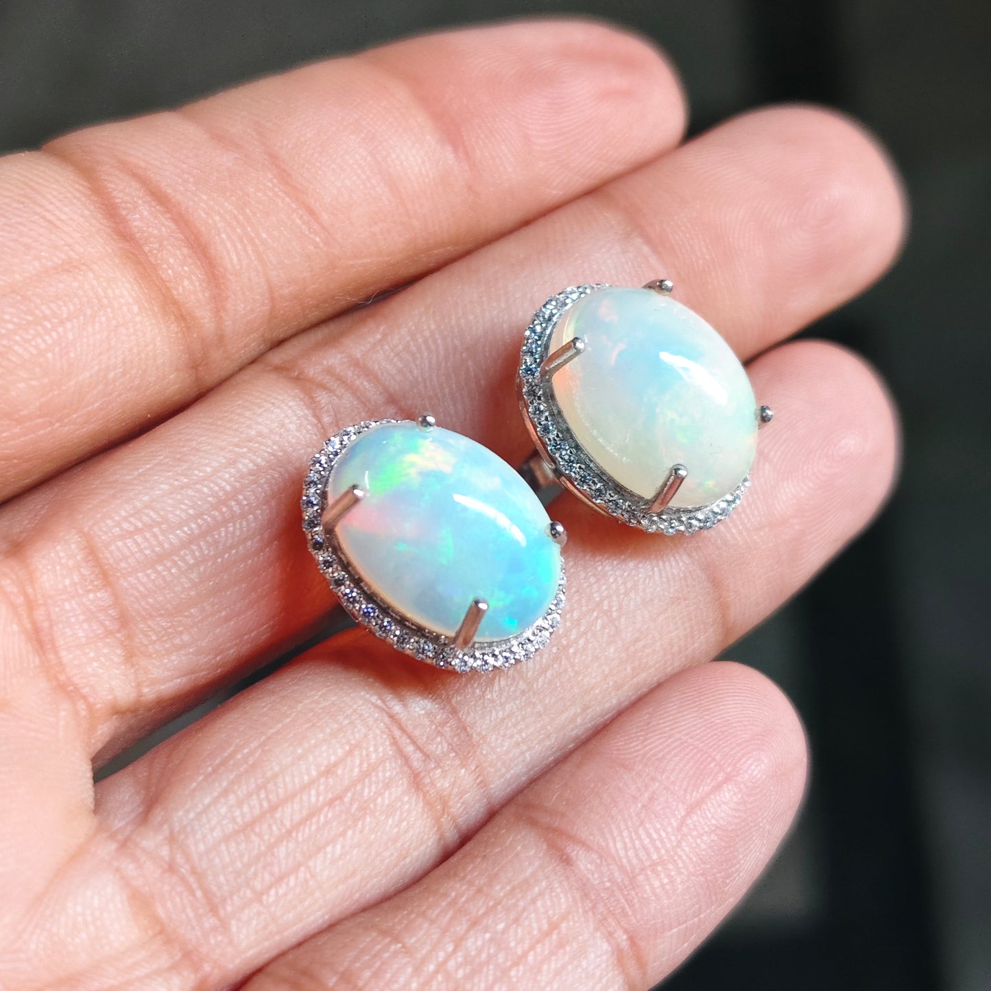 A pair of oval opal stud earrings with silver settings, held on a person's fingers. The opal stones show a milky white base with soft iridescent blue and green flashes.