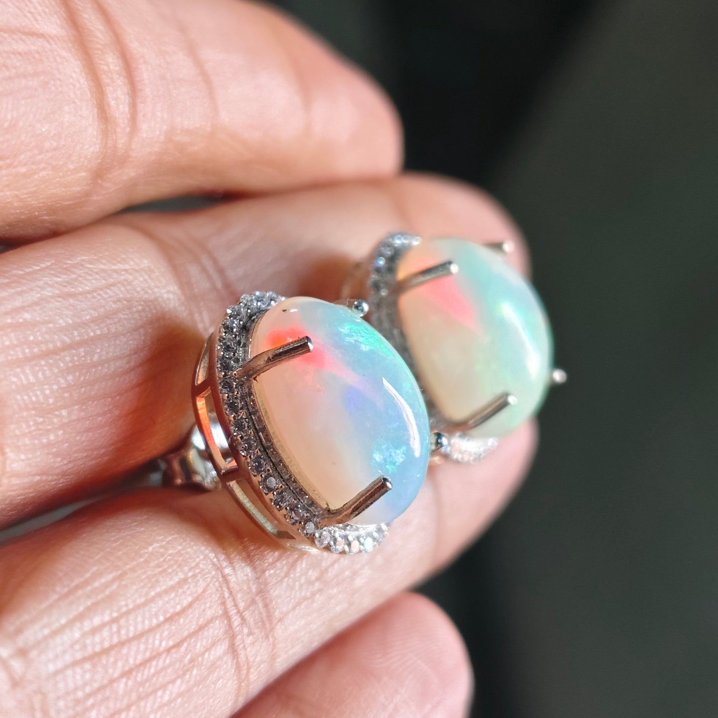 A close-up of a hand holding two oval opal rings with silver halos and small clear accent stones.