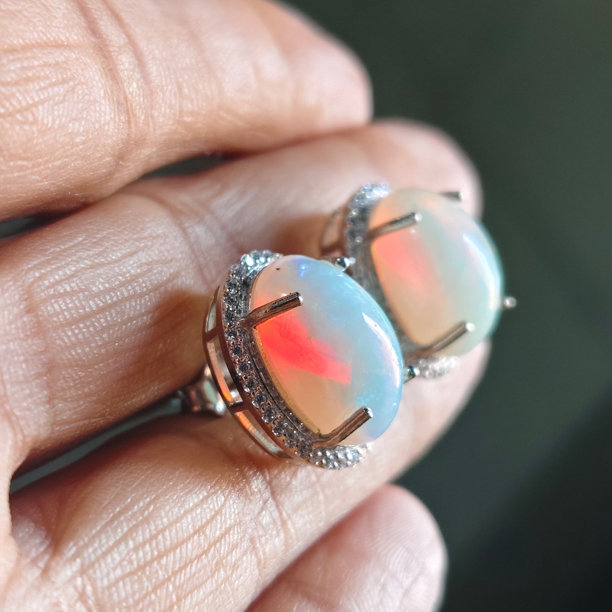 A close-up of a hand holding a pair of silver-toned ring settings with oval iridescent opal stones showing blue, white, and pink flashes.