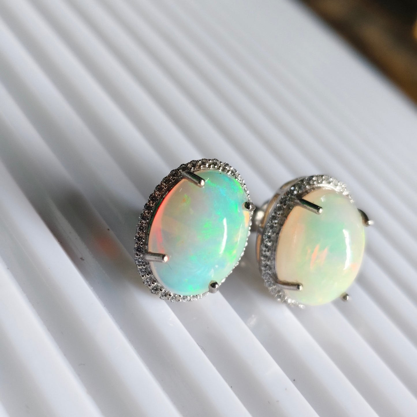 A pair of oval opal stud earrings in silver settings, resting on a white ribbed surface. The opal stones exhibit a play-of-color with iridescent hues of green, blue, and pink.
