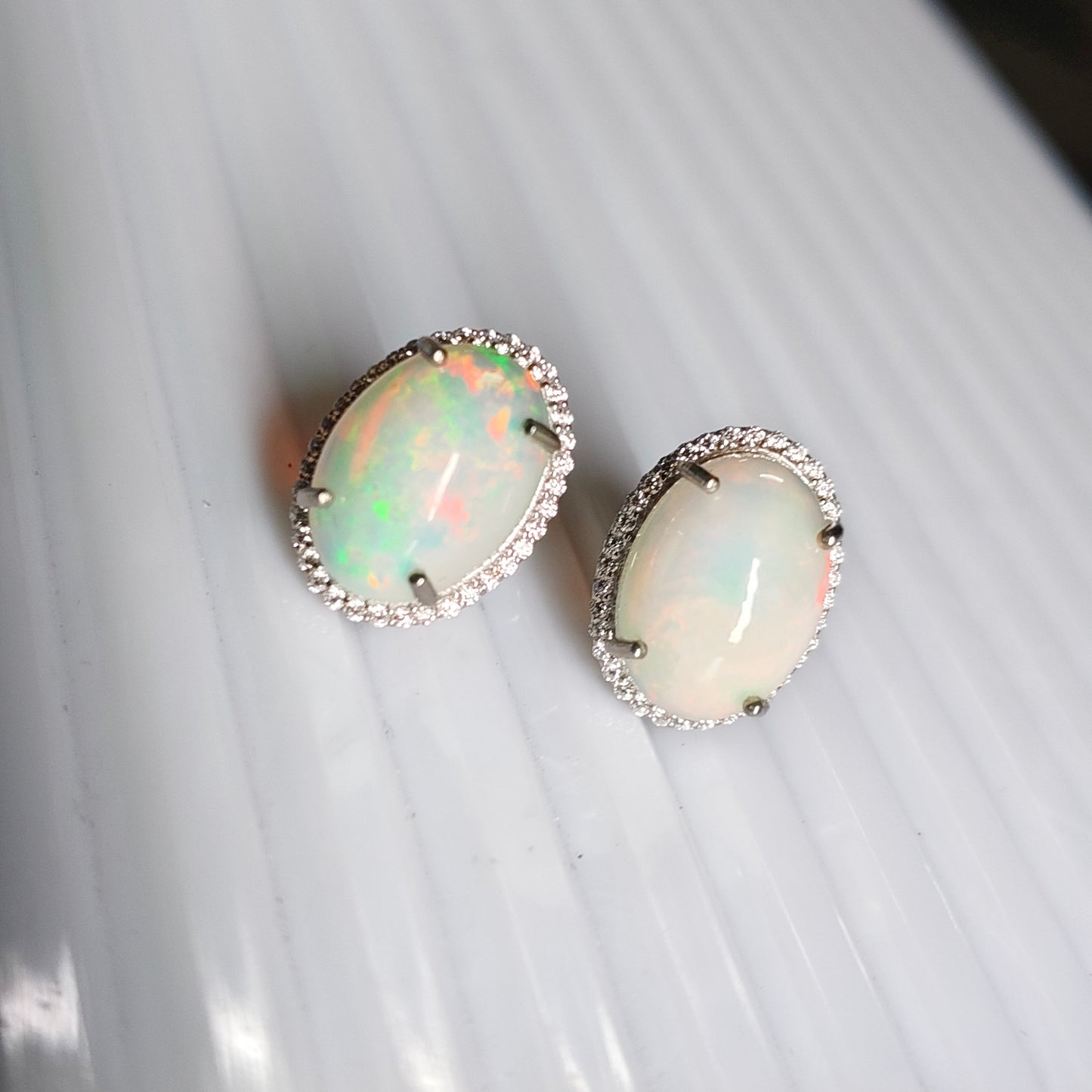 A pair of oval stud earrings featuring iridescent white opal gemstones in silver settings with small clear stones, displayed on a white ribbed surface.