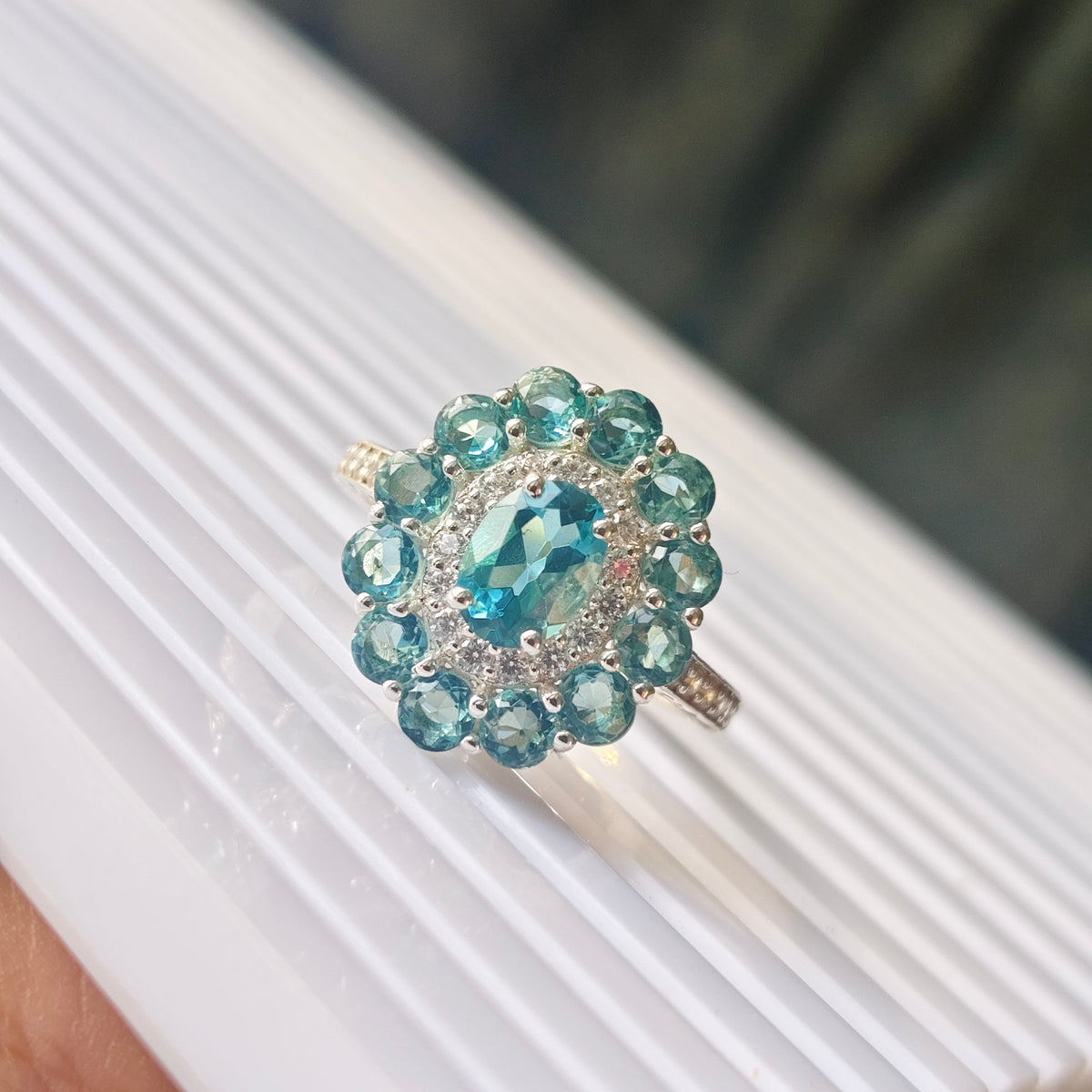A silver halo ring featuring a central oval-cut light blue gemstone surrounded by a double row of round-cut blue gemstones and small clear accent stones, displayed on a white ribbed surface.