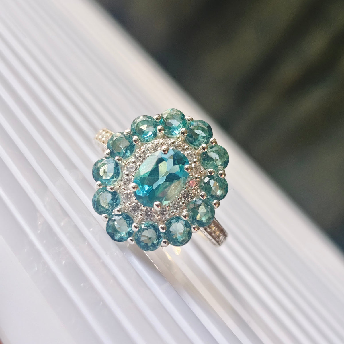 A silver ring featuring a large oval light blue gemstone surrounded by a halo of smaller round blue and clear stones, resting on a white ribbed surface.