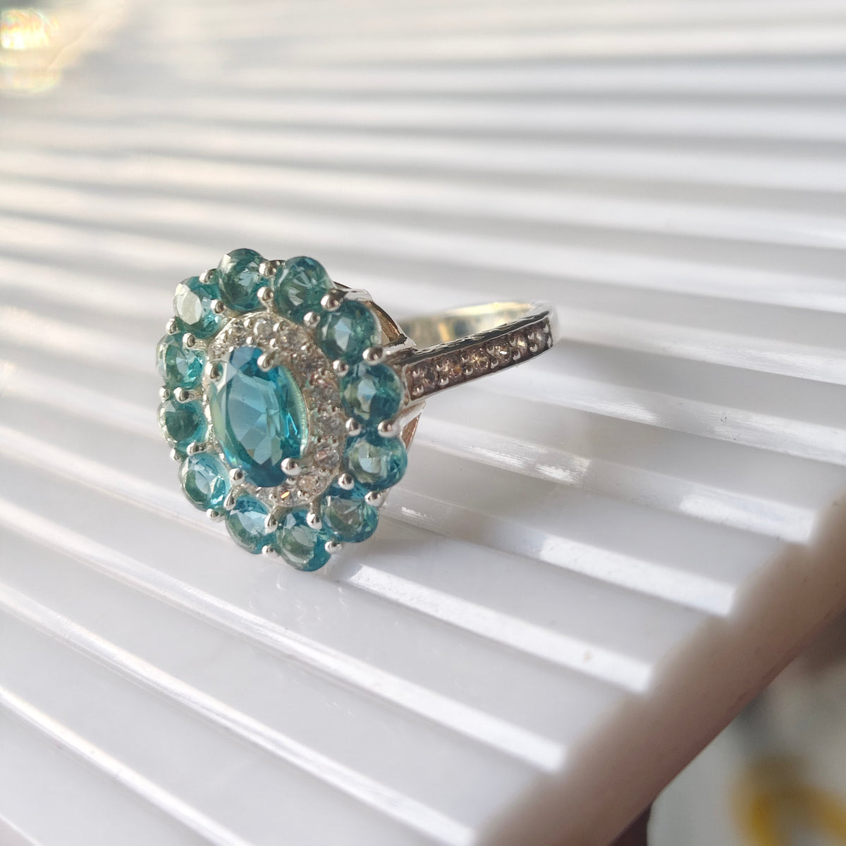 A silver-toned ring featuring a round cluster of light blue gemstones, including a central oval blue stone surrounded by smaller round stones, resting on a white ribbed surface.