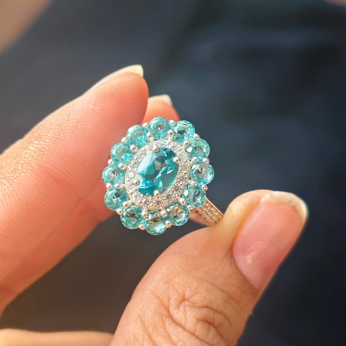A close-up of a hand holding a silver ring with a large oval-cut light blue center stone surrounded by a halo of small round blue stones and clear accent stones.