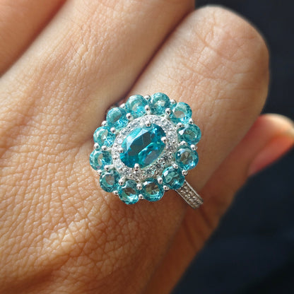 A close-up of a hand wearing a silver ring with a large oval-cut blue center stone surrounded by a halo of round blue stones and small clear accent stones.