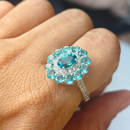 A close-up of a hand wearing a silver ring with a central light blue oval gemstone surrounded by a halo of small round blue and clear stones.
