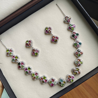 A jewelry set featuring a necklace and matching earrings with floral clusters of pink, green, and blue stones in silver settings, displayed inside an open black presentation box with a cream velvet lining.