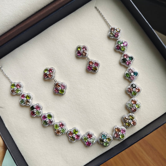 A jewelry set featuring a necklace and matching earrings with floral clusters of pink, green, and blue stones in silver settings, displayed inside an open black presentation box with a cream velvet lining.