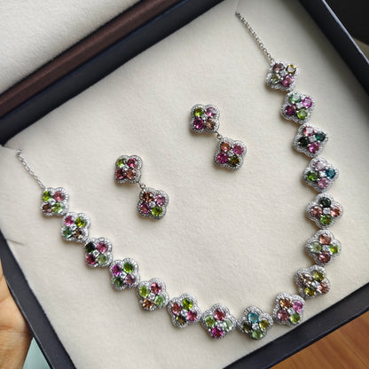 A silver-tone jewelry set with a necklace, bracelet, and matching earrings, each piece featuring multiple small round gemstones in pink, green, and blue, displayed inside an open black presentation box with a cream velvet lining.