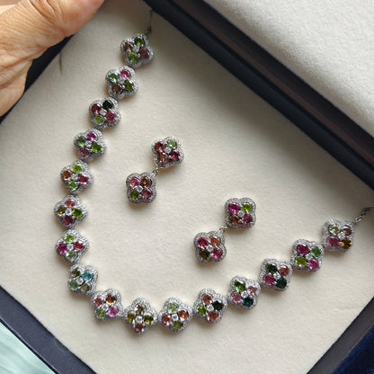 A jewelry set with a necklace and matching earrings displayed inside an open presentation box. The set features silver-toned metal with multiple colorful gemstones in pink, green, blue, and orange, arranged in a curved line on a cream-colored velvet insert.