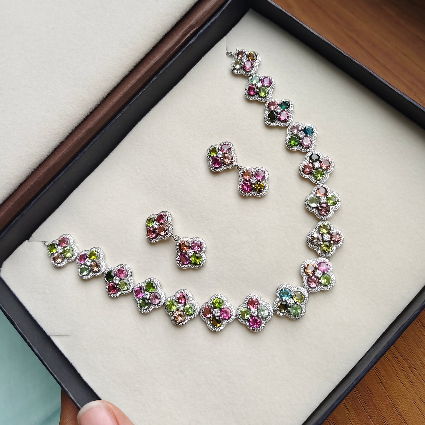 A jewelry set with a necklace and matching earrings displayed inside an open presentation box. The jewelry features small, colorful floral or heart-shaped clusters with pink, green, blue, and purple stones set in silver-toned metal.