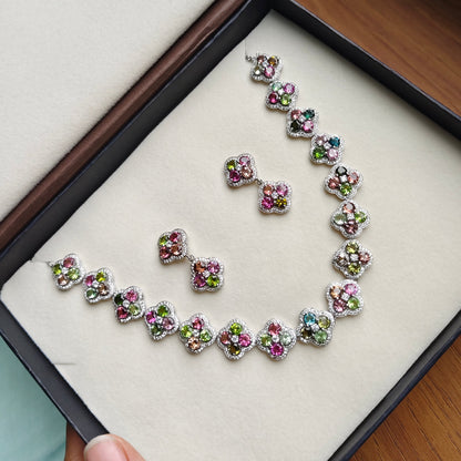 A jewelry set with a necklace and matching earrings displayed inside an open presentation box. The jewelry features small, colorful floral or heart-shaped clusters with pink, green, blue, and purple stones set in silver-toned metal.