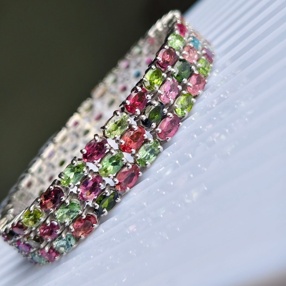 A close-up of a silver-toned bangle bracelet adorned with rows of oval-cut multicolor gemstones, including pink, green, and purple, resting on a white ribbed surface.