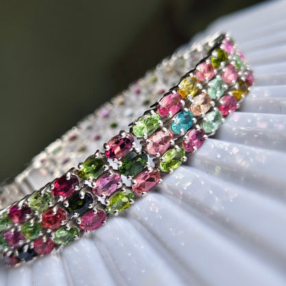 A close-up of a silver-toned cuff bracelet adorned with rows of colorful oval gemstones in pink, green, blue, yellow, and red, resting on a white pleated surface.