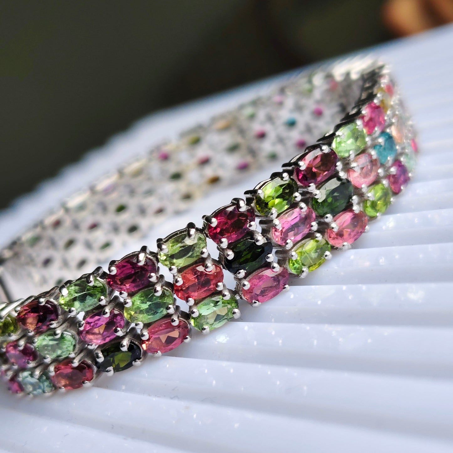 A close-up of a silver-toned bracelet adorned with multiple rows of colorful rectangular gemstones in pink, green, and blue tones, resting on a white ribbed surface.