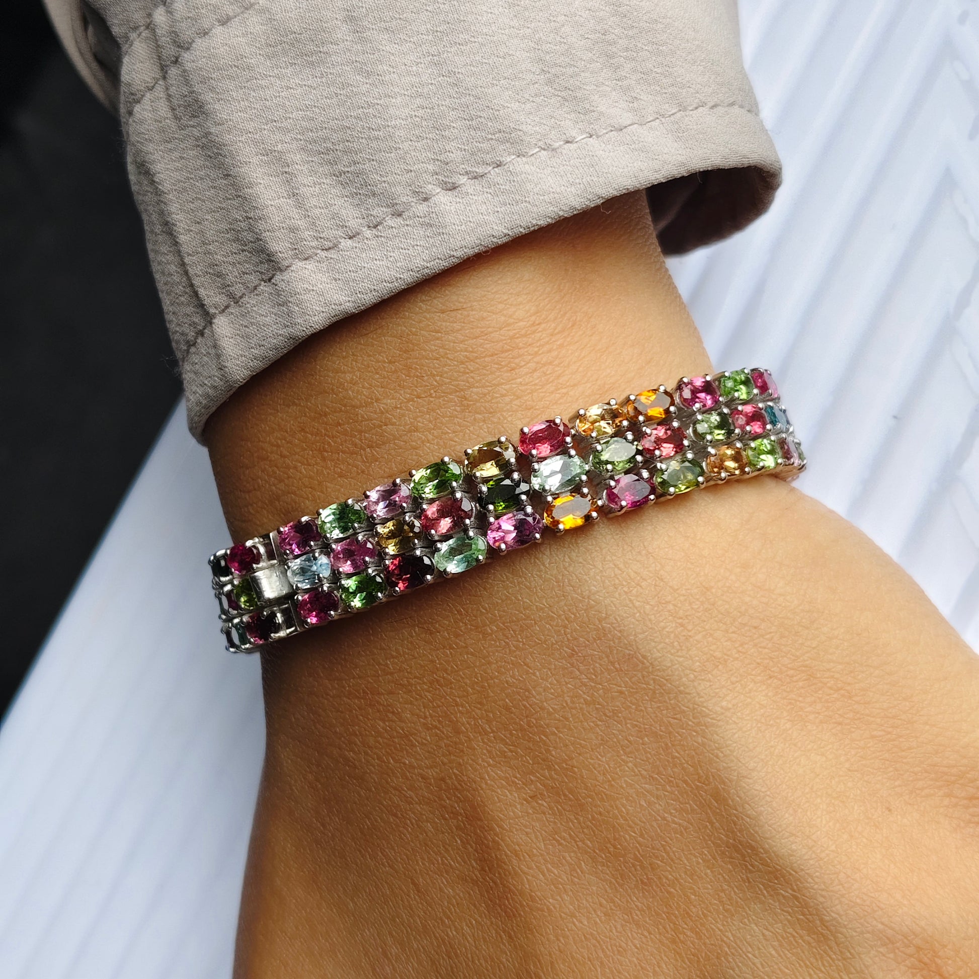 A close-up of a person's wrist wearing a multi-row bracelet made of colorful rectangular gemstones in pink, green, yellow, orange, and blue, paired with a light beige sleeve.