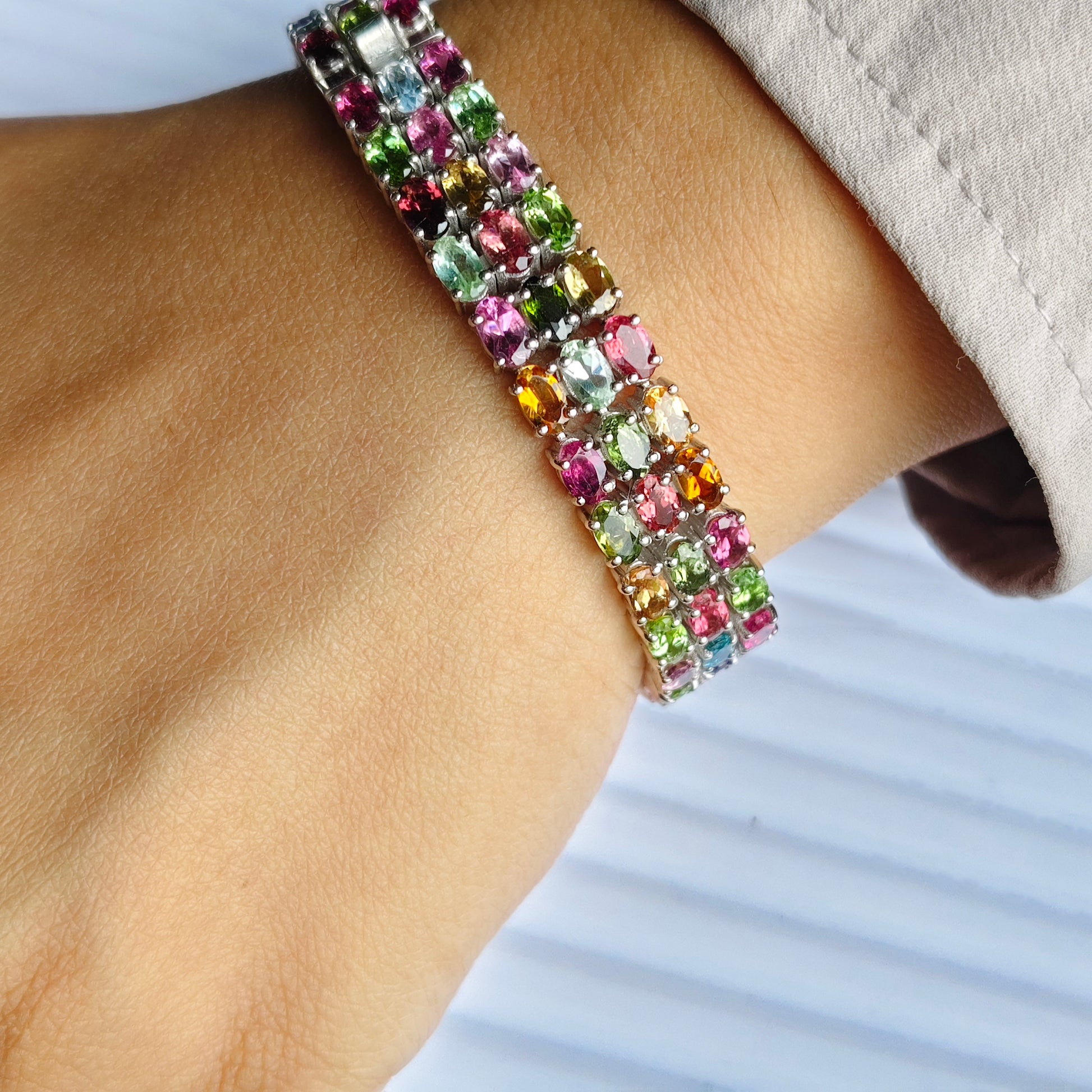 A close-up of a person's wrist wearing a multi-row bracelet set with colorful rectangular gemstones in pink, green, blue, yellow, and orange on a silver-toned band.