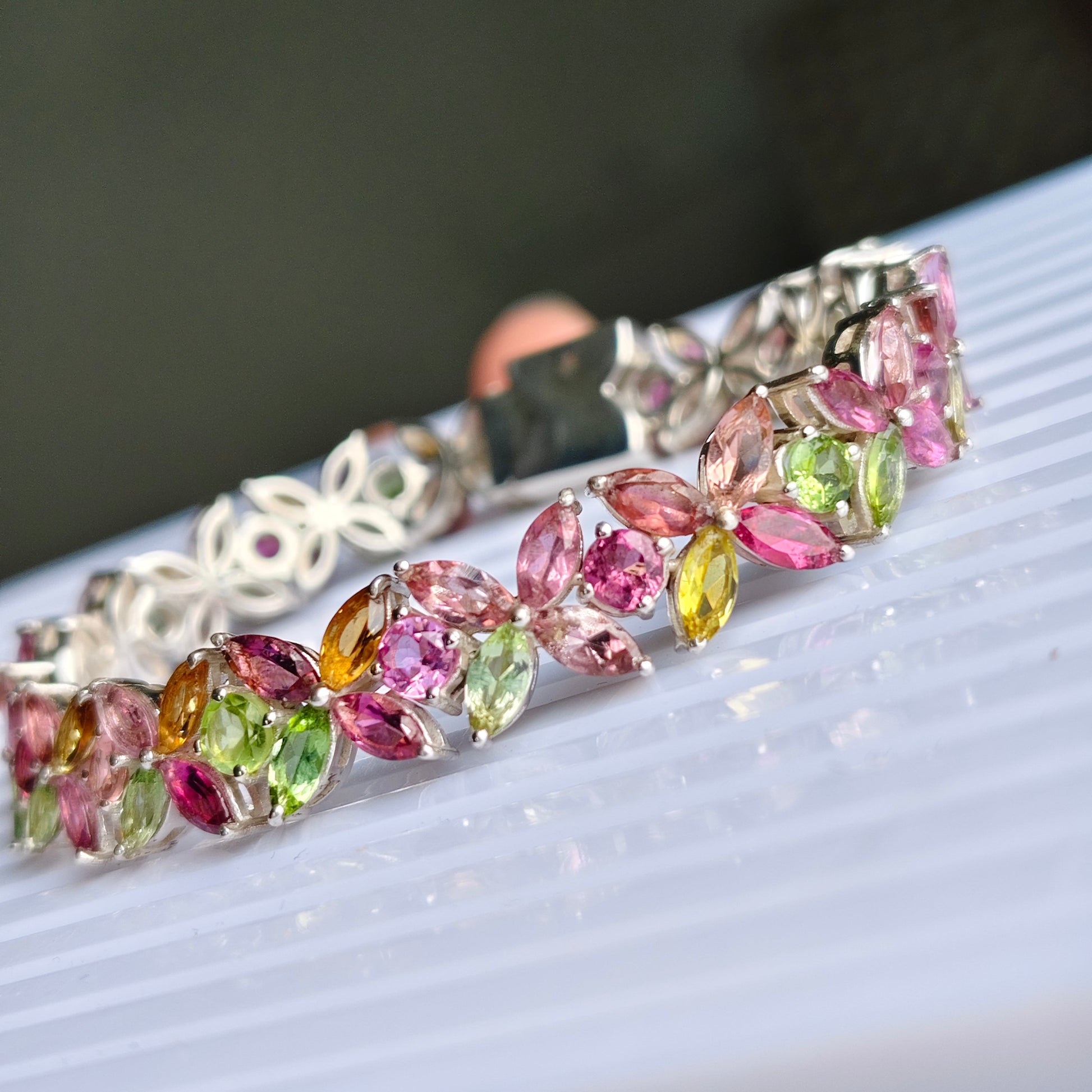 A close-up of a silver-toned bracelet adorned with marquise-cut multicolored gemstones in pink, green, yellow, and orange, resting on a white ribbed surface.