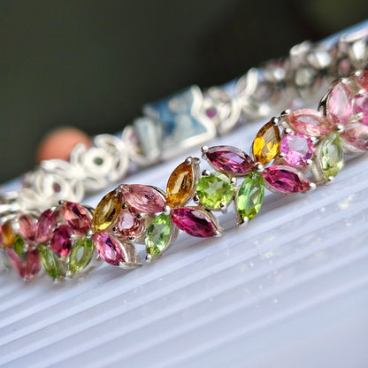 A close-up of a silver-toned bracelet adorned with marquise-cut gemstones in pink, green, yellow, and purple, resting on a white ribbed surface.
