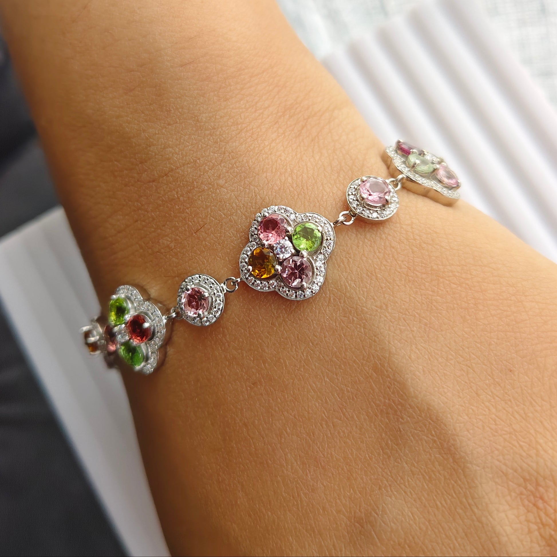 A close-up of a person's wrist wearing a silver-toned bracelet adorned with colorful gemstones, including pink, green, and orange stones in floral clover-like motifs.