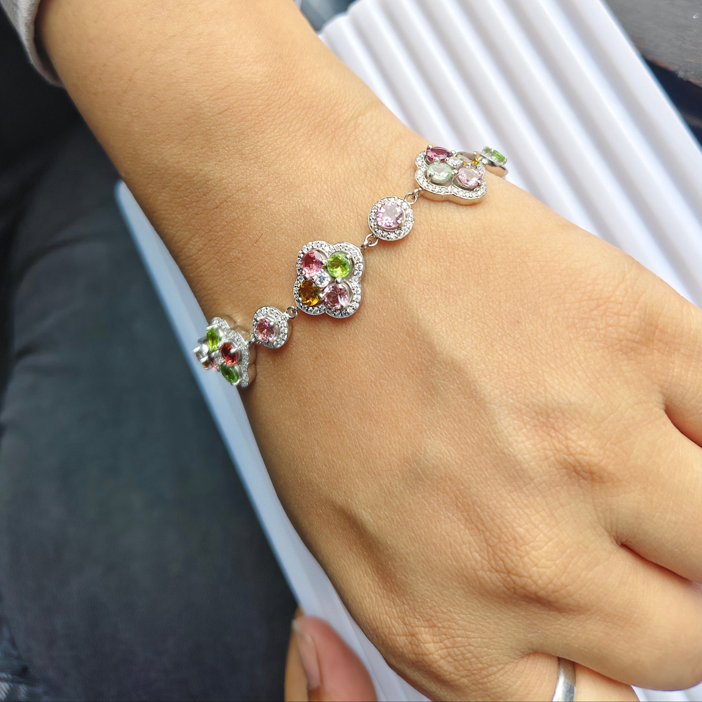 A close-up of a person's wrist wearing a silver-toned bracelet adorned with colorful gemstones, including pink, green, and purple accents, alongside a silver bangle.