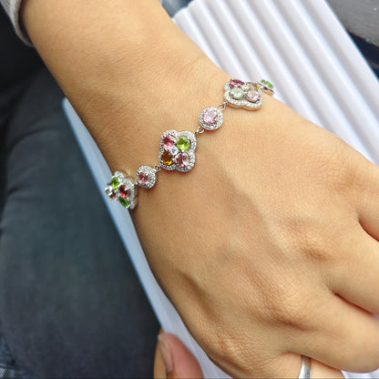 A close-up of a person's wrist wearing a silver-toned bracelet adorned with colorful gemstones, including pink, green, and purple accents, alongside a silver bangle.
