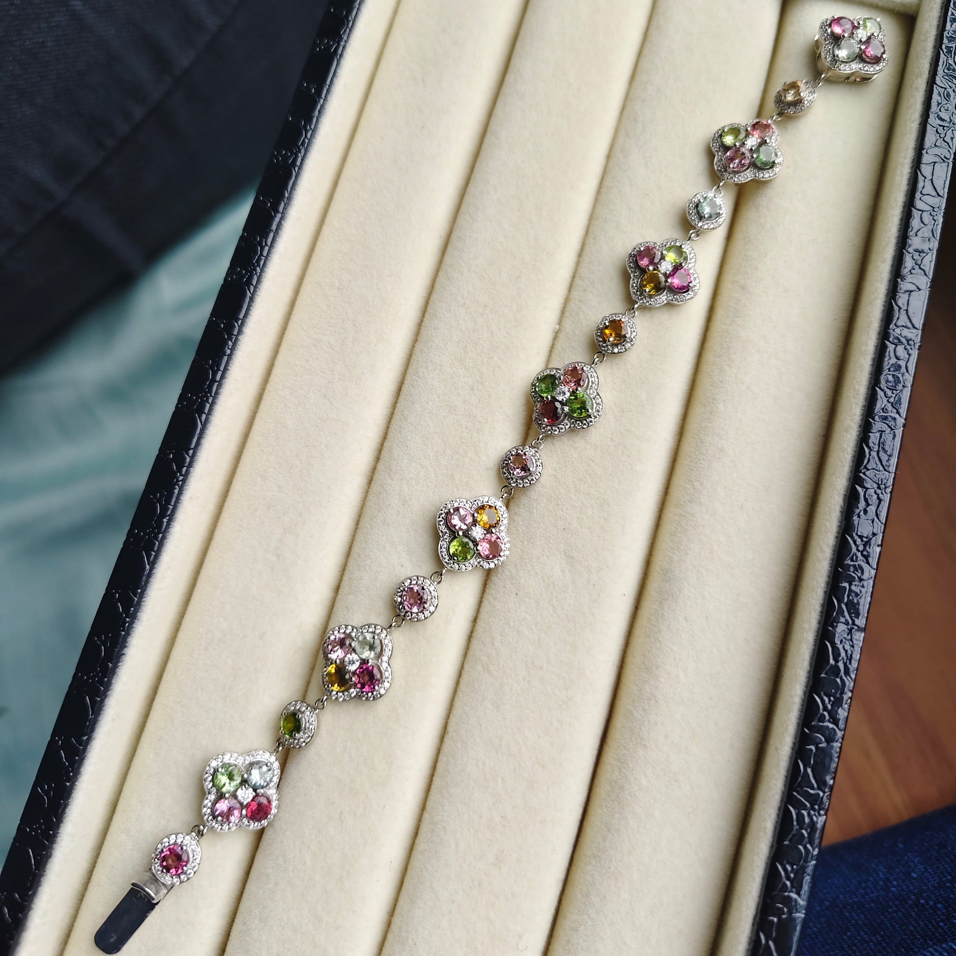 A delicate silver-toned bracelet with floral clusters of pink, green, and clear stones, displayed inside an open cream-colored jewelry box.